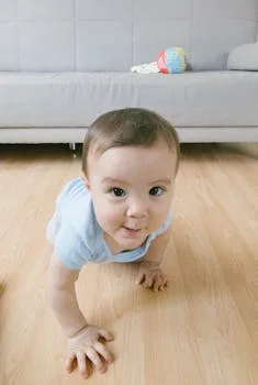 A cute baby crawling indoors on a wooden floor, looking at the camera.