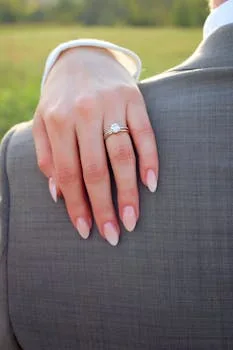 Close-up of a bride's hand with an elegant engagement ring on a groom's suit in a summer outdoor setting.